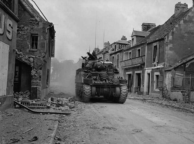 Photographie en noir et blanc – Les bâtiments d’une rue sont lourdement endommagés. Un char d’assaut Sherman se fraie un chemin à travers les décombres.